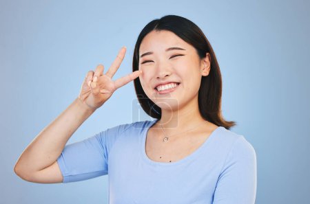 Photo for Peace, sign and portrait of woman with hand for emoji in studio blue background with gen z style, fashion or happiness. Face, smile and Asian model with gesture, expression. - Royalty Free Image