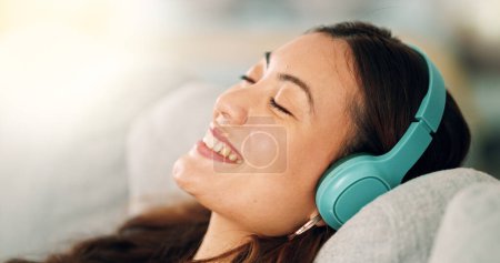 Téléchargez les photos : Détendez-vous, écouteurs et femme heureuse écoutant de la musique, radio ou podcast tout en se reposant sur le canapé. Bonheur, sourire et fille calme de singapore assis sur le canapé dans le salon à la maison avec la technologie. - en image libre de droit