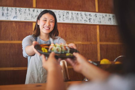 Téléchargez les photos : Sushi, travailleuse de restaurant et femme heureuse avec service de nourriture et repas asiatique dans une cuisine. Assiette, sourire de serveur féminin au travail ou chef travaillant avec la recette de poisson pour le déjeuner commande avec cuisine au bar japonais. - en image libre de droit