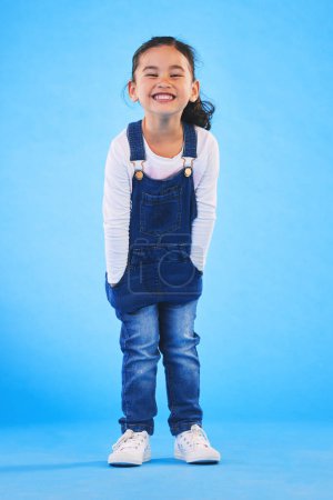 Photo for Excited girl child, fashion and studio portrait with jeans, dungaree and trendy style by blue background. Female kid model, smile and happy for clothes, aesthetic and youth culture by backdrop. - Royalty Free Image