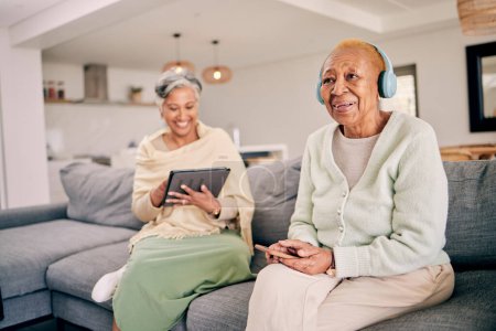 Téléchargez les photos : Écouteurs, relaxer et les femmes âgées sur le canapé à la maison écouter de la musique, audio et radio avec un ami. Retraite, technologie et personnes dans le salon sur tablette pour le streaming piste, chanson ou abonnement. - en image libre de droit