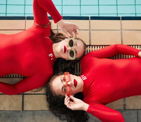 Téléchargez les photos : Mode, lunettes de soleil et maillots de bain de femmes amies en body rouge à la piscine intérieure. Portrait au-dessus des jeunes femmes avec style sportif, élégance et art ou luxe dans des vêtements de créateurs. - en image libre de droit