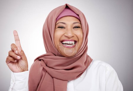 Foto de Señalando hacia arriba, retrato y una mujer musulmana en un fondo de estudio con una sonrisa para una pregunta. Feliz, gesto y una persona árabe o dama islámica con un dedo para un voto aislado en un telón de fondo. - Imagen libre de derechos