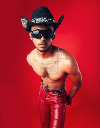 Photo for Fashion, man and cowboy in hat, shirtless with sunglasses and gen z isolated on red background. Confident model, cool and leather pants with eyewear accessory, western aesthetic and edgy in studio. - Royalty Free Image