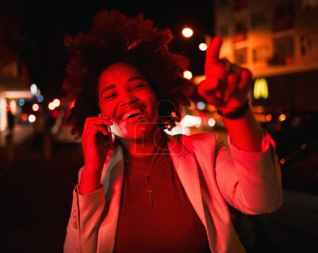 Téléchargez les photos : Femme d'affaires, appel téléphonique dans la rue et les lumières de la ville dans la nuit, sourire et pointant du doigt en attendant le taxi. Smartphone, éclairage rouge néon et heureuse femme noire sur la route avec conversation en réseau - en image libre de droit