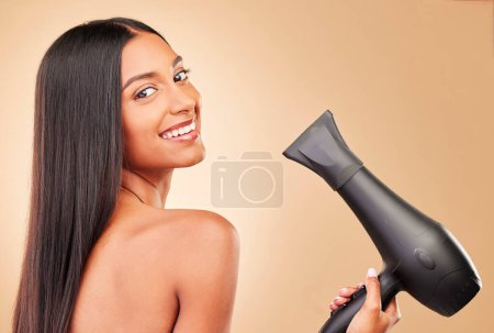 Téléchargez les photos : Sèche-cheveux, sourire et portrait de femme en studio avec coiffure de salon propre pour le bien-être. Santé, bien-être et heureux modèle féminin avec outil cosmétique pour l'entretien par un fond brun - en image libre de droit