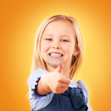 Photo for Child, happy portrait and thumbs up in studio for support, like emoji or yes for approval. Face of young girl kid on a orange background for hand gesture, icon or sign for agreement or thank you. - Royalty Free Image