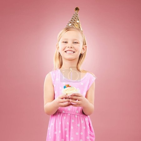 Cupcake, cumpleaños y retrato de un niño con un sombrero para la fiesta de vacaciones o la celebración feliz. Muchacha emocionada sobre un fondo rosa para sorpresa, pastel o celebrar el logro con un postre y alegría.