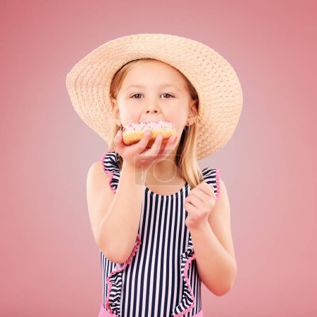 Photo for Girl kid, eating a donut and dessert, sweets and summer with hat and bathing suit isolated on pink background. Portrait, youth and cake with snack, child enjoying bakery treat with hat in studio. - Royalty Free Image