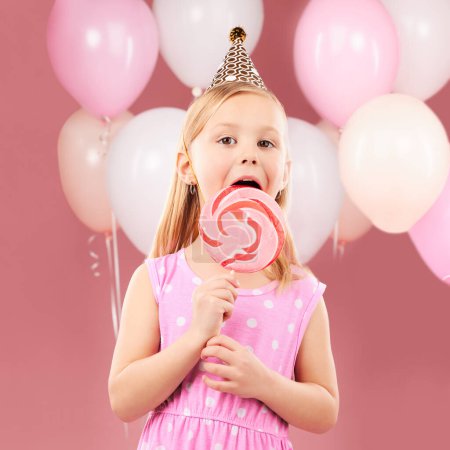 Téléchargez les photos : Ballons, sucette et portrait d'enfant sur fond rose pour fête d'anniversaire, célébration et journée spéciale. Joyeux, excité et jeune fille mignonne sourire avec des bonbons, friandises sucrées et dessert en studio. - en image libre de droit
