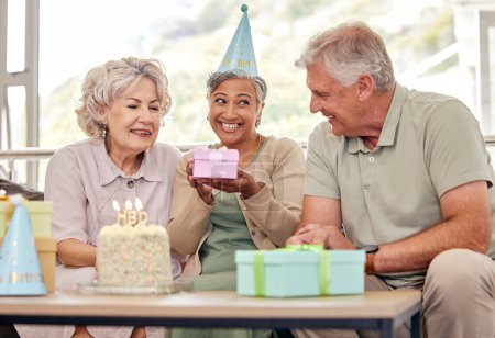 Foto de Cumpleaños, amigos mayores con pastel y regalo para la celebración en la sala de estar, sonrisa y velas en la jubilación. Felicidad, ancianos y fiesta con postre, sorpresa y anciano hombre con mujeres en sofá - Imagen libre de derechos