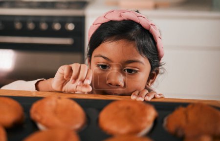 Photo for Baking, muffin and a sneaky indian girl in the kitchen of her home to steal a fresh pastry from the counter. Food, children or cooking with a young kid looking naughty at a baked snack closeup. - Royalty Free Image