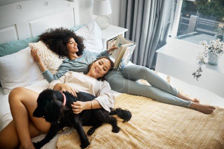Photo for Dog, bed and happy lesbian couple in home, morning and relax together reading book. Pet, bedroom and gay women with animal, bonding and play in healthy relationship, love connection and interracial. - Royalty Free Image