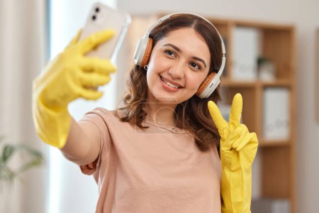 Photo for Cleaner woman, selfie and peace sign with smile, headphones and audio streaming for music in home. Happy maid girl, emoji or icon with photography, memory and profile picture on social network app. - Royalty Free Image