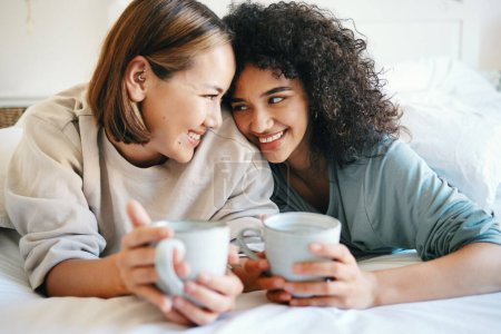 Photo for Coffee, love and lesbian couple on bed in conversation for bonding and relaxing together. Happy, rest and interracial lgbtq women laughing, talking and drinking latte in bedroom of modern apartment - Royalty Free Image