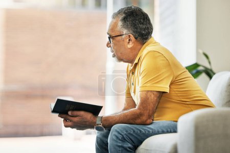 Photo for Senior man, thinking and reading a book on sofa in home living room learning knowledge, information or ideas. Elderly, person and relax in retirement with novel, books or inspiration for fiction. - Royalty Free Image