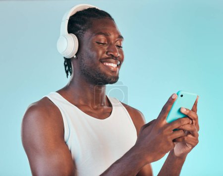 Photo for Headphones, cellphone and black man in a studio networking and listening to music, playlist or album. Happy, smile and African male model scroll on internet with phone and song by blue background - Royalty Free Image