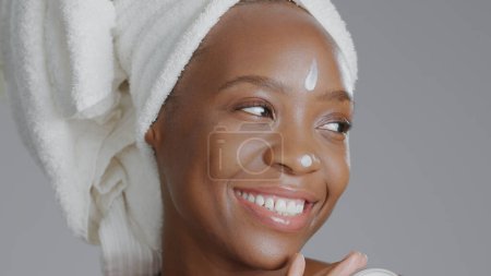 Photo for Face, beauty and cream with a happy black woman in studio on a gray background for natural wellness. Skincare, smile and lotion with a happy young model closeup to apply treatment to her skin. - Royalty Free Image