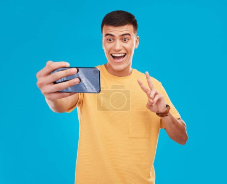 Téléchargez les photos : Homme, selfie et paix signe de la main avec bonheur, excité par les médias sociaux avec communication sur fond bleu. V emoji, influenceur de mémoire et sourire en photo avec photographie dans un studio. - en image libre de droit