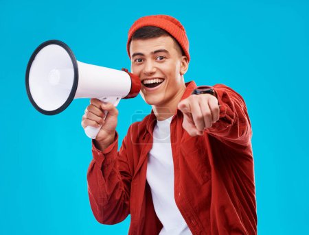 Photo for Portrait, bullhorn and young man in studio pointing for an announcement or speech at a rally. Happy, smile and male activist on stool with megaphone for loud communication isolated by blue background. - Royalty Free Image