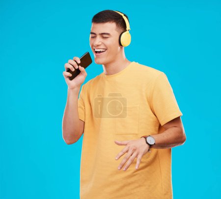 Photo for Phone, headphones and man singing in studio listening to music, radio or playlist for entertainment. Smile, technology and male person streaming song or album for karaoke isolated by blue background - Royalty Free Image