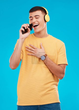 Photo for Man, headphones and singing to music with smartphone, listening to radio with karaoke on blue background. Fun, playful and entertainment with technology, audio streaming and using phone app in studio. - Royalty Free Image