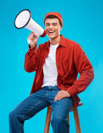 Photo for Portrait, megaphone and young man in a studio for an announcement or speech for a rally. Happy, smile and male activist on stool with bullhorn for loud communication isolated by blue background - Royalty Free Image