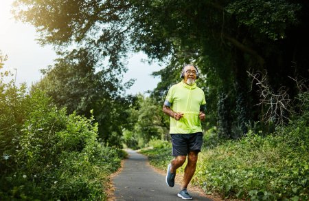 Photo for Senior man, running and listening to music on road in nature, countryside or forest for exercise in retirement. Fitness, training and workout or run on path in woods for cardio, health and wellness. - Royalty Free Image