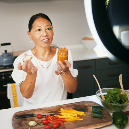 Photo for Asian woman, vegetables and live streaming for cooking with bottle, healthy food and blog on social media in kitchen. Chef, mature influencer and content creation, class and broadcast online tutorial. - Royalty Free Image