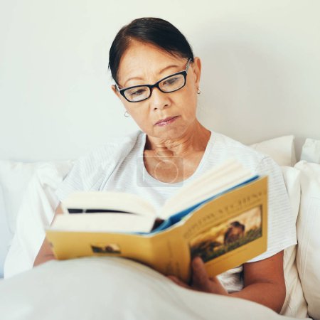 Photo for Mature woman reading books in bedroom for story, fiction novel and knowledge at home. Serious asian person, glasses and comfortable in bed with literature for rest, hobby and relax to enjoy break. - Royalty Free Image
