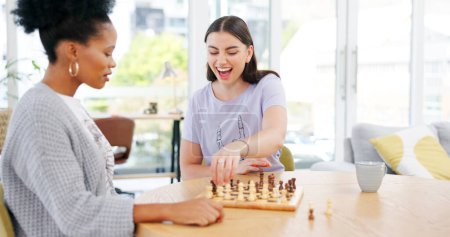 Téléchargez les photos : Filles amies jouant aux échecs dans le salon pour se lier, se divertir ou s'amuser ensemble. Joyeux, sourire et les jeunes femmes interracial profiter du jeu de société dans le salon de l'appartement moderne - en image libre de droit