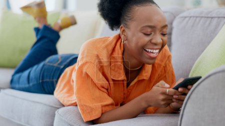 Téléchargez les photos : Détendez-vous, mème de téléphone drôle ou femme noire sur le canapé en communication dans le salon de la maison. Sourire, défiler ou dame africaine heureuse sur le canapé rire, envoyer des SMS ou lire des commérages en ligne sur la discussion de l'application mobile. - en image libre de droit