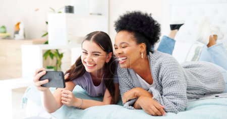 Photo for Happy, selfie and girl friends on a bed for bonding and relaxing together with a phone. Smile, technology and interracial female people taking a picture with cellphone in bedroom of modern apartment - Royalty Free Image