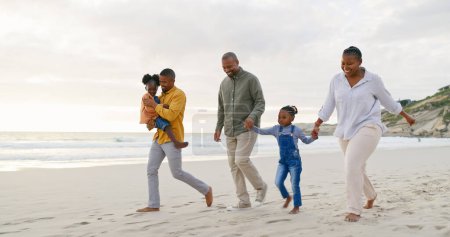Foto de Feliz, caminando y una familia negra en la playa, tomados de la mano y hablando en unas vacaciones. Puesta de sol, conversación y abuelos, padre e hijos en un paseo por el mar durante unas vacaciones para viajar. - Imagen libre de derechos