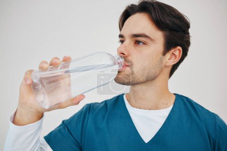 Photo for Nurse, man drinking water in bottle and health, wellness or body nutrition in studio isolated on a white background in hospital. Medical professional, hydration and liquid of thirsty surgeon on break. - Royalty Free Image