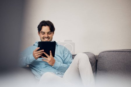 Foto de Hombre feliz, tableta y relajarse en el sofá para las redes sociales, la comunicación o el entretenimiento en la sala de estar en casa. Hombre persona, sonrisa y acostado en el sofá salón con la tecnología para la transmisión en línea en casa. - Imagen libre de derechos