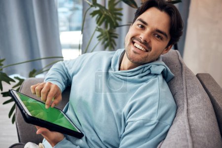 Foto de Retrato, tableta y pantalla verde con un hombre jugando en un sofá en la sala de estar de su casa para relajarse. Tecnología, sonrisa y joven jugador feliz usando una pantalla con marcadores de seguimiento en su apartamento. - Imagen libre de derechos