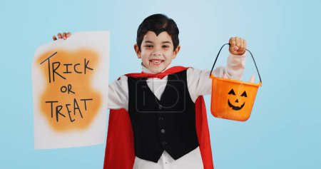 Téléchargez les photos : Halloween, affiche et portrait d'enfant avec panier de bonbons pour bonbons isolé en studio fond bleu et heureux. Vacances, rires et enfants en costume de vampire pour la célébration dans le festival de fantaisie. - en image libre de droit
