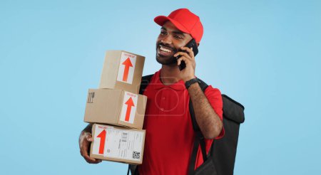Photo for Man, logistics and courier with cellphone in communication to client, delivery or boxes in studio. Indian person, supply chain and cargo in conversation with customer, packages and blue background. - Royalty Free Image