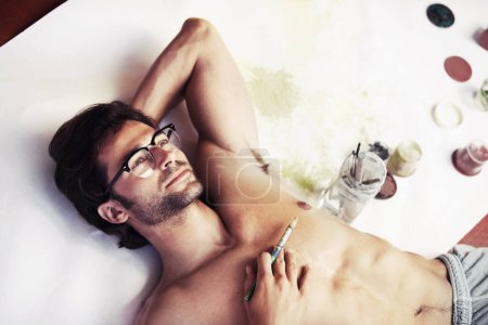 Photo for Artist, lying and thinking with paintbrush for relaxing in space with canvas, paints or jars for work. Young man, face and glasses for inspiration of creative, project or hobby by shirtless in room. - Royalty Free Image