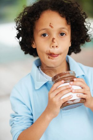 Photo for Cute, portrait and child with chocolate spread at a home with delicious, sweet snack or treat. Smile, happy and face of young boy kid from Mexico eating nutella jar licking lips at modern house - Royalty Free Image