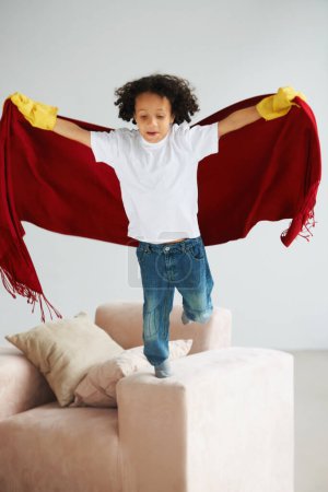 Photo for Fly, children and a boy superhero in a costume on a sofa in the living room of his home for playing a game. Jumping, growth and fantasy with a young kid in a cape for justice or safety cosplay. - Royalty Free Image