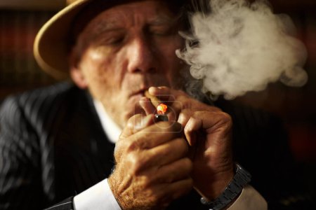 Foto de Maduro, hombre y cigarrillo con encendedor para fumar para relajarse en el espacio. Personas mayores, jefe de la mafia o gángster con traje, sombrero y cara seria por hábito, mano y soplo para reunirse en actividad delictiva. - Imagen libre de derechos