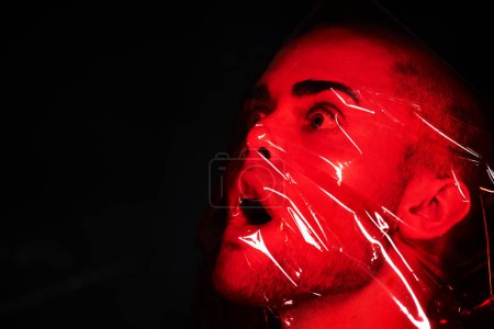 Foto de Hombre, plástico rojo y sofocado en un estudio aislado sobre una maqueta de fondo negro. Polietileno, película y persona con depresión, miedo y ansiedad, estrés y crisis de salud mental, psicología y neón. - Imagen libre de derechos