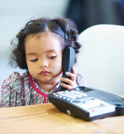 Foto de Escuchar, hablar y un niño en una llamada telefónica para comunicarse en una casa. Inicio, contacto y una niña, niño o bebé hablando por teléfono fijo para conversar, jugar o discutir en un escritorio. - Imagen libre de derechos