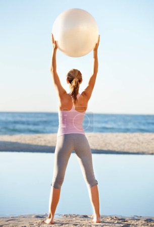 Téléchargez les photos : Femme, tenir et pilates ballon pour la forme physique sur la plage en étirant pour l'équilibre dans le bien-être avec vue arrière. Femme, yoga et mouvement dans la nature pour la paix, calme ou zen dans la préparation à l'entraînement. - en image libre de droit