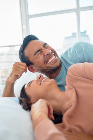 Photo for Happy young mixed race couple in a loving relationship laughing while relaxing together in bed at home. Boyfriend making a silly face while telling funny and lighthearted jokes to cheerful girlfriend. - Royalty Free Image