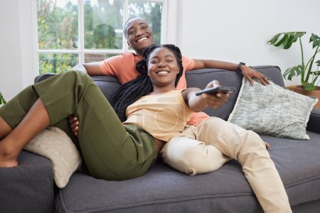 Photo for Young african american couple changing channels on remote and watching television together on sofa at home. Girlfriend relaxing on boyfriends lap while enjoying entertainment shows, series and movies. - Royalty Free Image