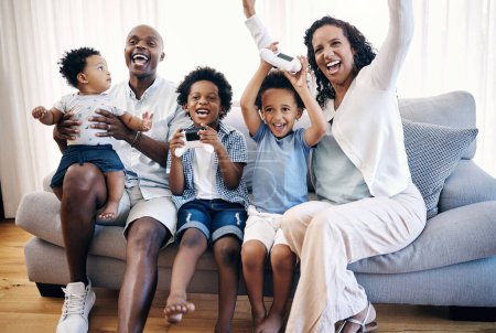 Téléchargez les photos : Heureuse jeune famille afro-américaine assise sur un canapé dans le salon à la maison et célébrant une victoire. Adorables garçons métis jouant à des jeux vidéo avec des consoles pendant que leurs parents les encouragent. - en image libre de droit