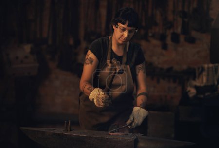Foto de El trabajo duro siempre es el camino. una joven golpeando una barra de metal caliente con un martillo en una fundición - Imagen libre de derechos
